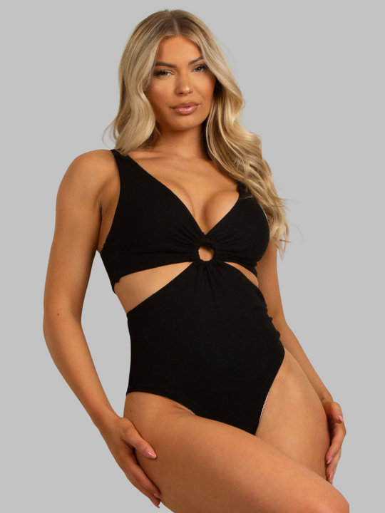 Female model wears a black ribbed cut out bodysuit with v neck design and ruched centre detailing. The model stands in front of a plain light blue background. The model has tanned skin and long blonde hair, she faces the camera and places her hands by her sides.