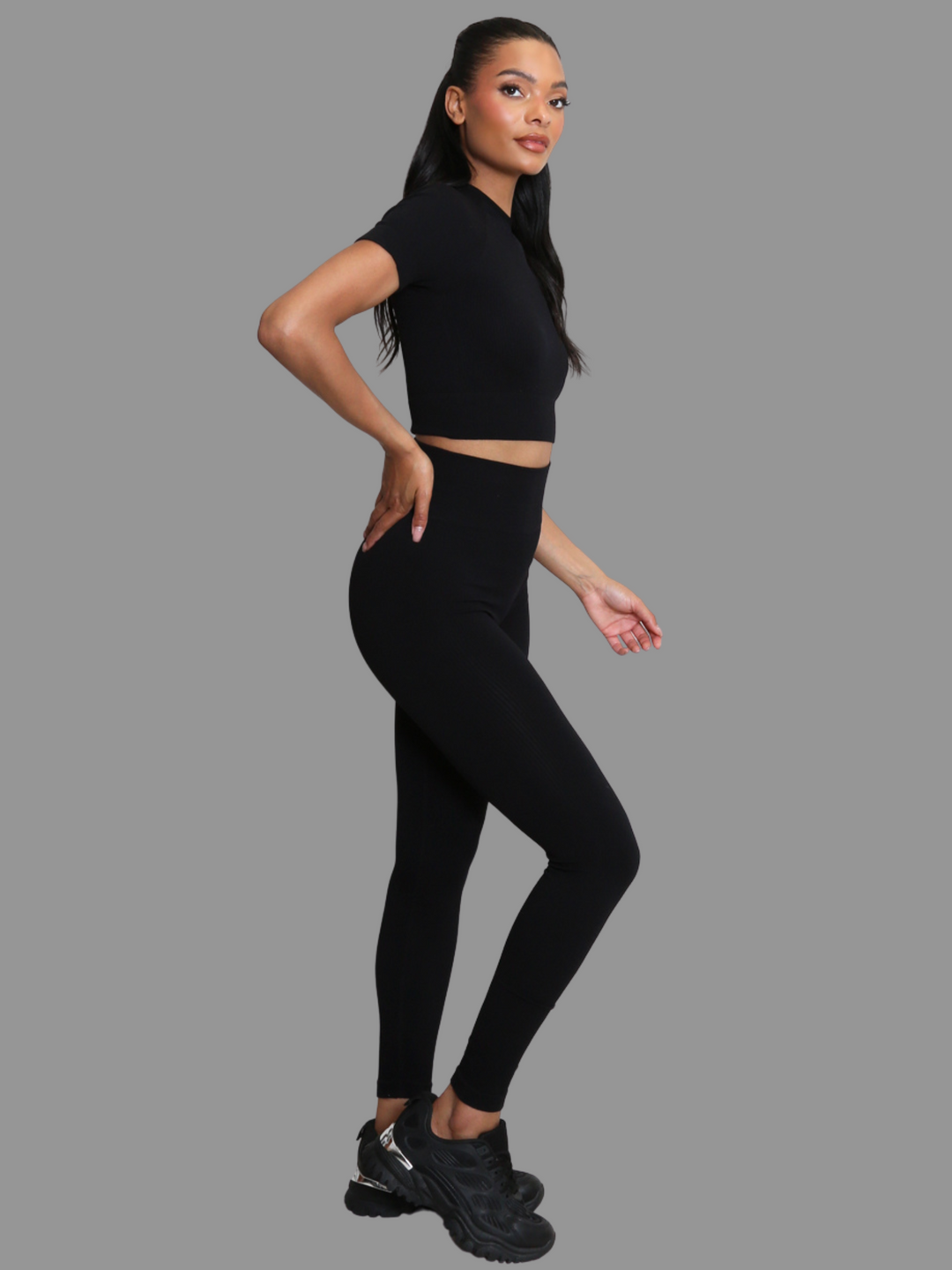Full length shot. female model stands in front of a grey background. Model wears a black ribbed co-ordinated set that has a black ribbed crop top with short sleeves and matching leggings. The model has light brown skin and long black hair, she stands to the side and places one hand on her hip, the side of the co-ordinated set can be seen. 