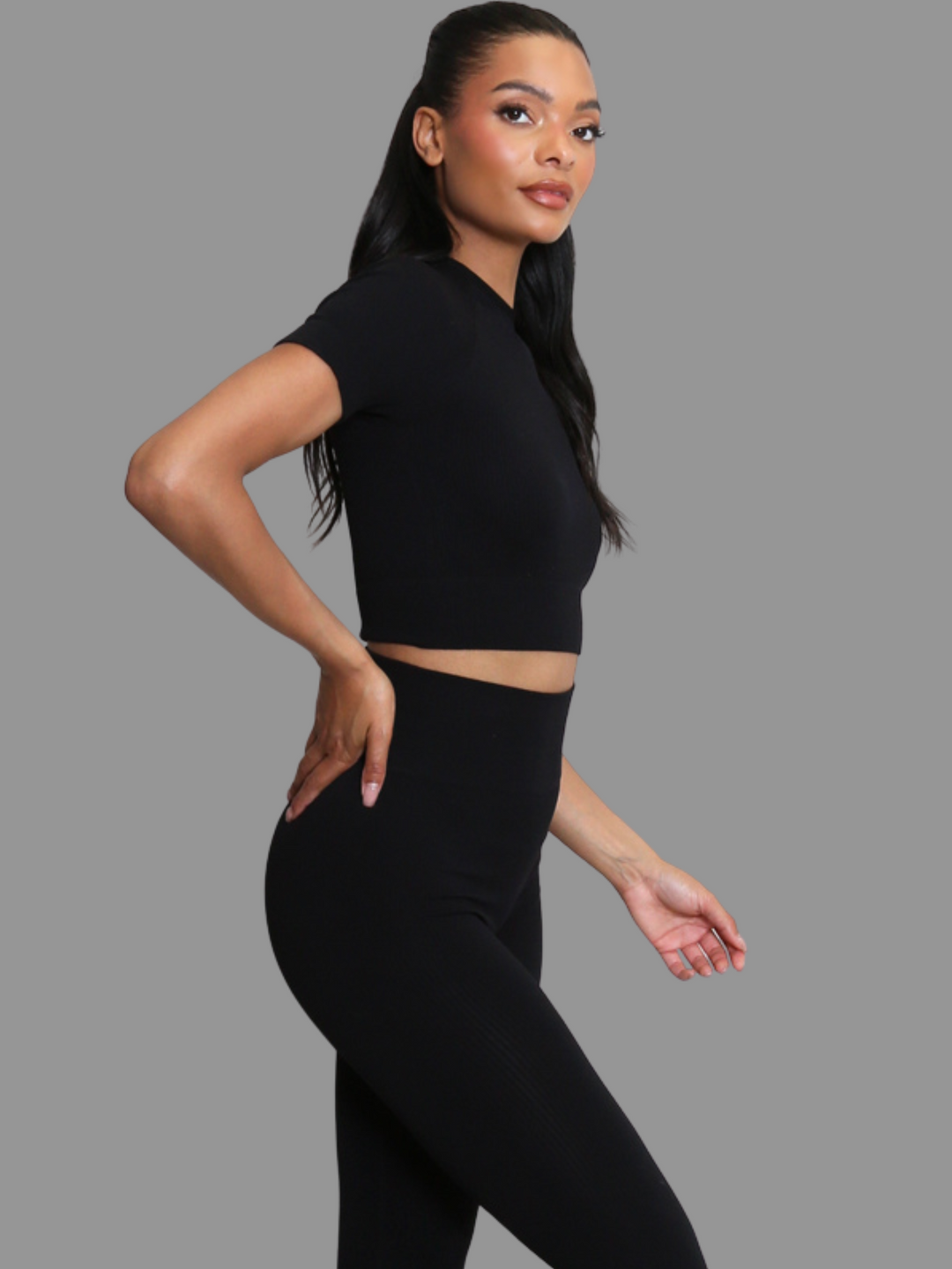 female model stands in front of a grey background. Model wears a black ribbed co-ordinated set that has a black ribbed crop top with short sleeves and matching leggings. The model has light brown skin and long black hair, she stands to the ide and places one hamd on her hip, she turns her head to look into the camera. 