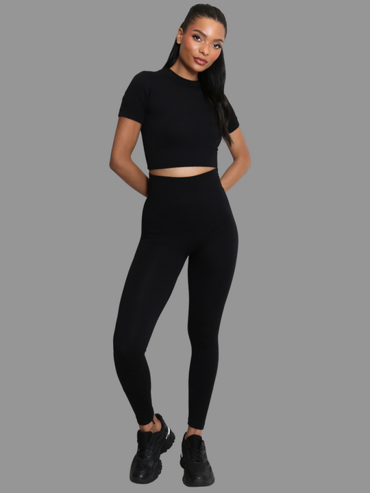 Full length shot. female model stands in front of a grey background. Model wears a black ribbed co-ordinated set that has a black ribbed crop top with short sleeves and matching leggings. The model has light brown skin and long black hair, she faces the camera and places both hands behind her back. 