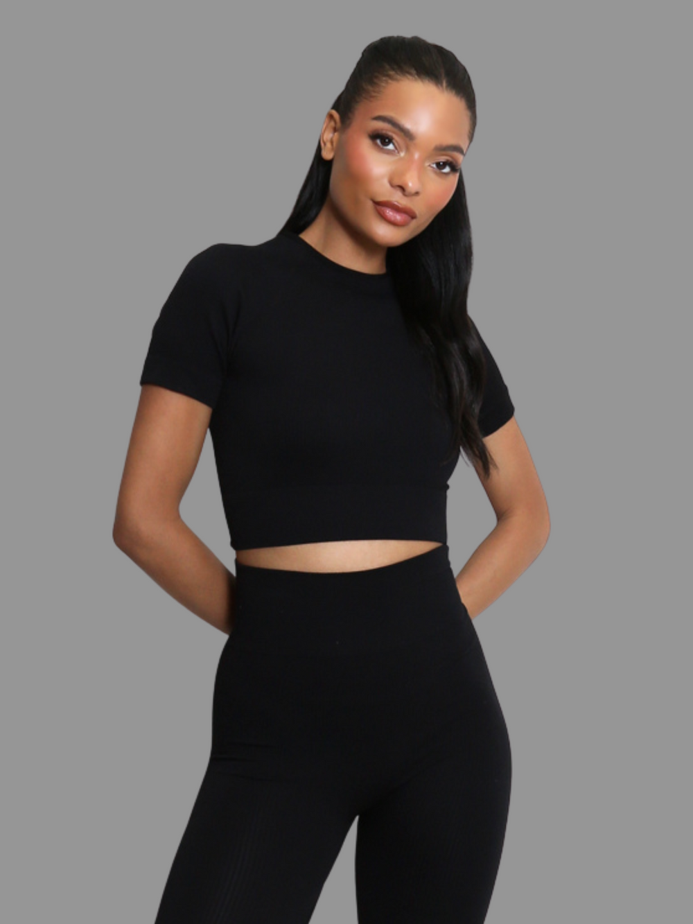 female model stands in front of a grey background. Model wears a black ribbed co-ordinated set that has a black ribbed crop top with short sleeves and matching leggings. The model has light brown skin and long black hair, she stands facing forward and looks directly into the camera. The model places her hands behind her back. 