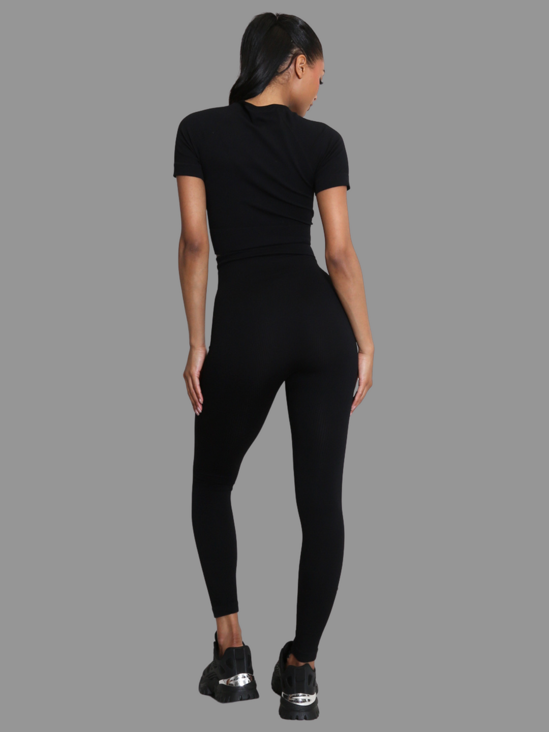 Full length shot. female model stands in front of a grey background. Model wears a black ribbed co-ordinated set that has a black ribbed crop top with short sleeves and matching leggings. The model has light brown skin and long black hair, she stands with her back to the camera, the back of the co-ordinated set can be seen.