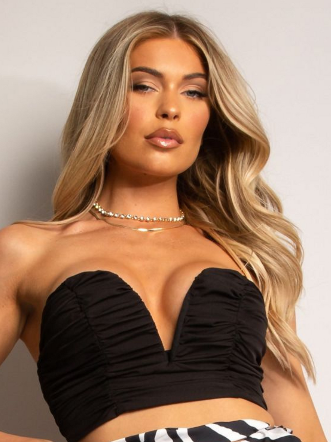 Model wears a black bralette with ruched design and a plunging sweatheart neckline. The black bralette top has a zip back fastening. The model has blonde hair, she looks directly into the camera, the v neck sweatheart cut of the top can be seen.