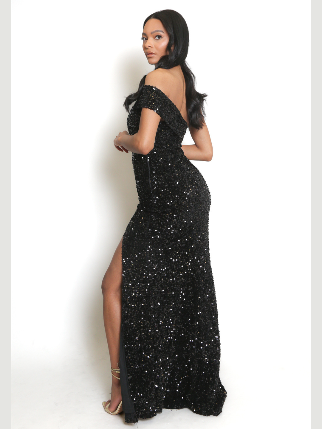 Full length shot. Female model stands in front of a plain background. The model wears a black sequin maxi dress with front side split and off the shoulder sequinned sleeve. The model has brown skin and long black hair, she stands to the side, the side split can be seen. 