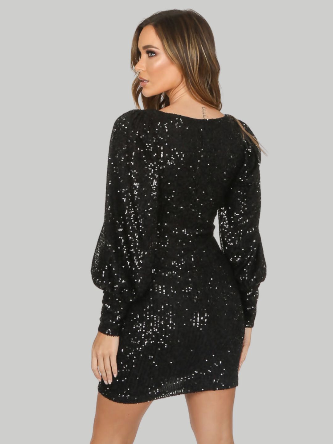 Female model wears a v neck black sequin midi dress, with puffed long sleeves. The model has tanned skin and light brown hair, she stands with her back to the camera and places her hands by her side, she looks to the side.