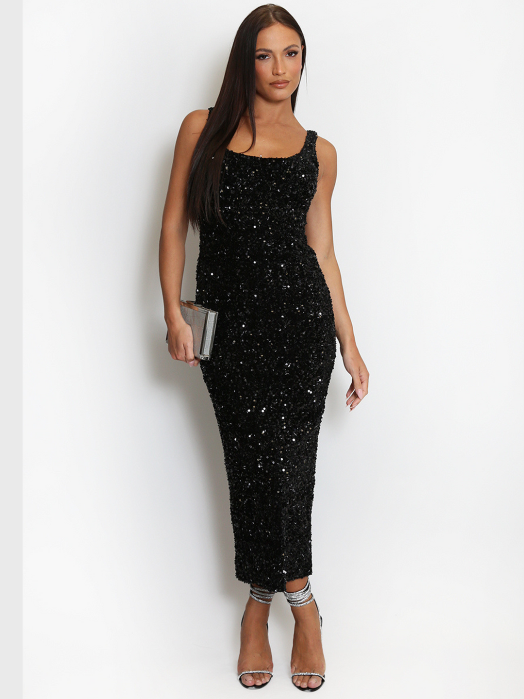 Model weas a  deluxe black sequin midi dress with a square neckline and a sleeveless cut. The model has tanned skin and long brown hair. The model faces the camera, the front of the sequin dress can be seen, she wears a strappy diamante heel and crosses over her feet, she holds a silver clutch bag in one hand and her hand by her side. 