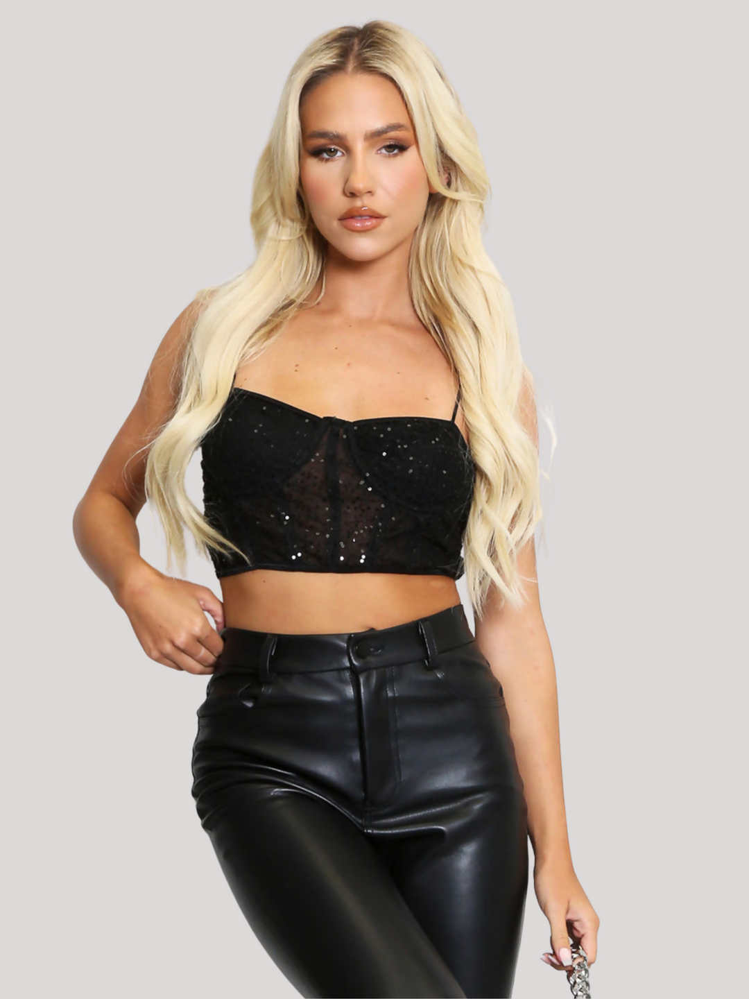 Female model stands before a plain backround. The model wears a black sequin crop top with hook back fastening and spaghetti straps and black pu leather trousers. The crop top has a front sheer panel with sequin detailing. The model has tanned skin and long blonde hair, she faces the camera, placing one hand by her side and the other at her hip. 