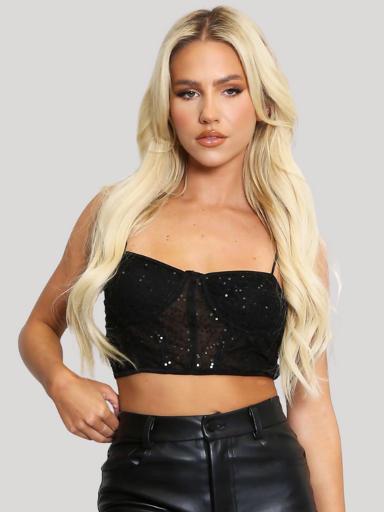 Female model stands before a plain backround. The model wears a black sequin crop top with hook back fastening and spaghetti straps. The crop top has a front sheer panel with sequin detailing. The model has tanned skin and long blonde hair, she faces the camera, placing one hand on her hip and the other by her side.
