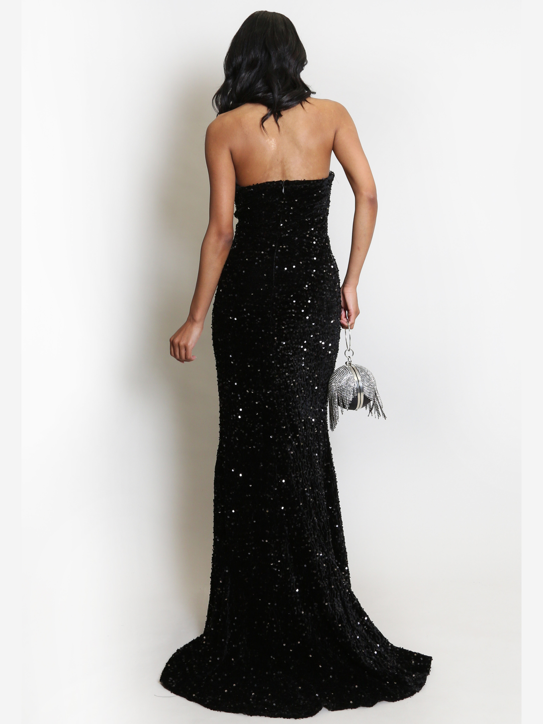 Full length shot. Model stands before a plain background. The model wears a black sequin maxi dress, with a fish tail hem and silver sequin sweetheart neckline. The model  has light brown skin and long black hair. The model stands with  her back to the camera, she holds  a Diamante bag in the other. 