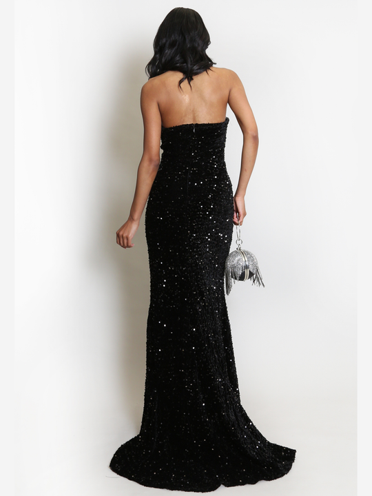 Full length shot. Model stands before a plain background. The model wears a black sequin maxi dress, with a fish tail hem and silver sequin sweetheart neckline. The model  has light brown skin and long black hair. The model stands with  her back to the camera, she holds  a Diamante bag in the other. 