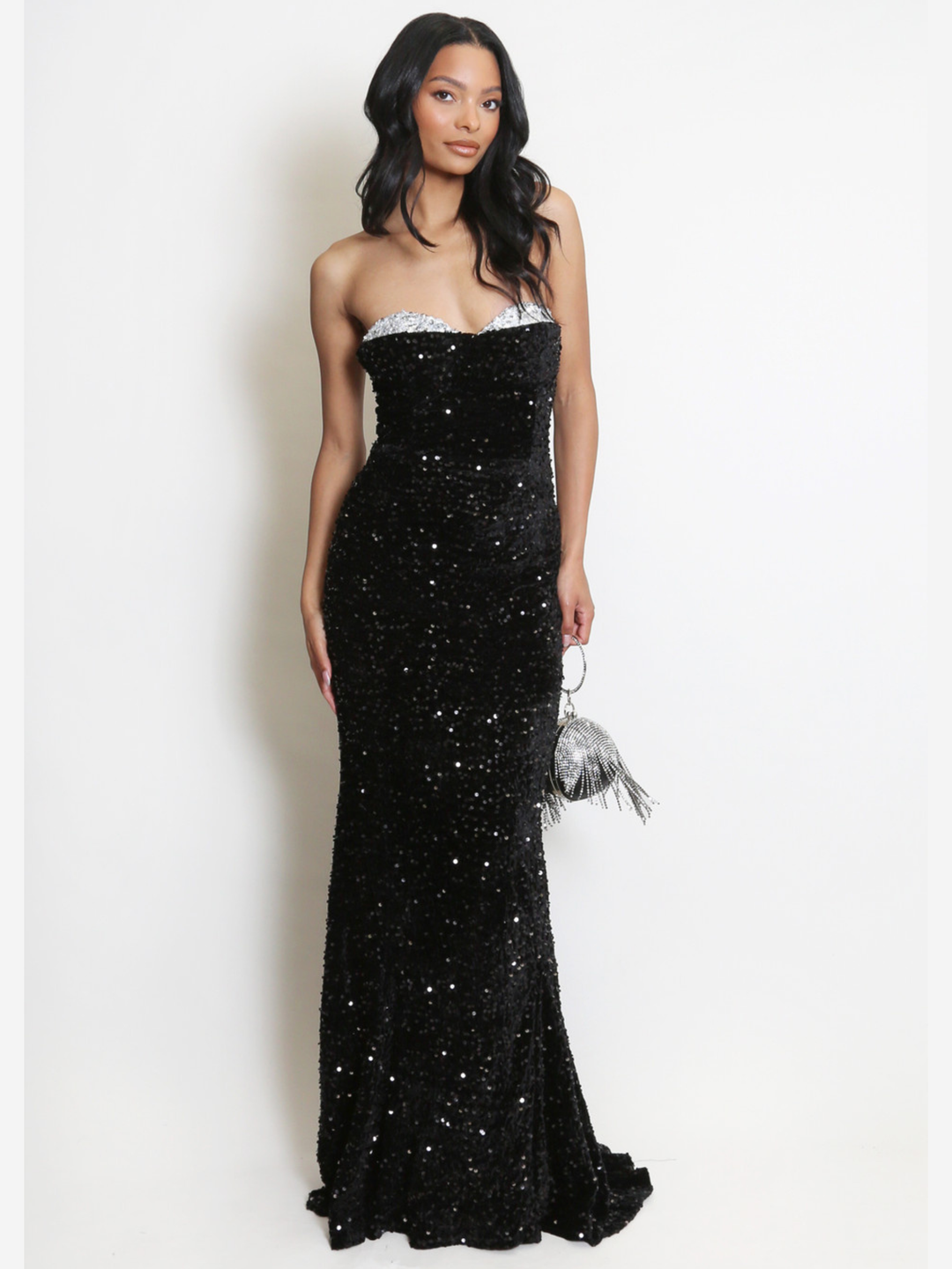 Full length shot. Model stands before a plain background. The model wears a black sequin maxi dress, with a fish tail hem and silver sequin sweetheart neckline. The model faces the camera, she has light brown skin and long black hair. The model places one hand by her side  and holds a Diamante tasseled bag in the other.  The sequinned fish tail trail can be seen.