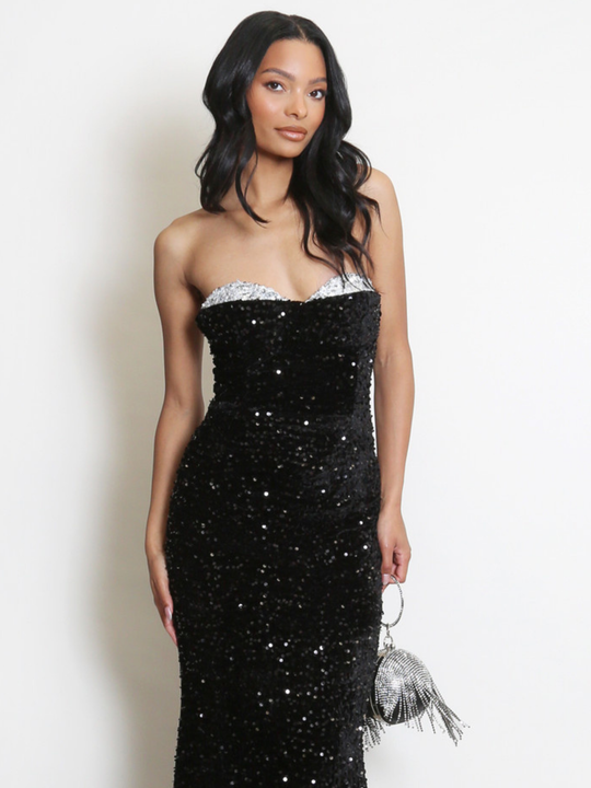 Model stands before a plain background. The model wears a black sequin maxi dress, with a fish tail hem and silver sequin sweetheart neckline. The model has light brown skin and long black hair. The model faces the camera and holds a Diamante hand bag. 