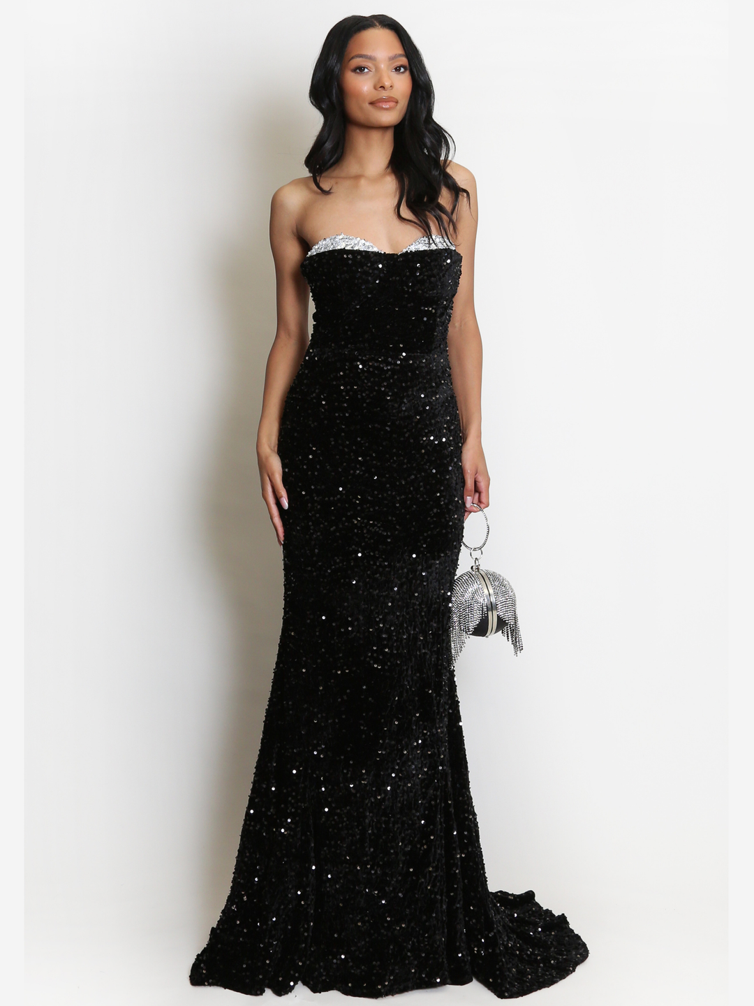 Full length shot. Model stands before a plain background. The model wears a black sequin maxi dress, with a fish tail hem and silver sequin sweetheart neckline. The model has light brown skin and long black hair. The model faces forward, she holds a Diamante bag  and her hands by her sides.