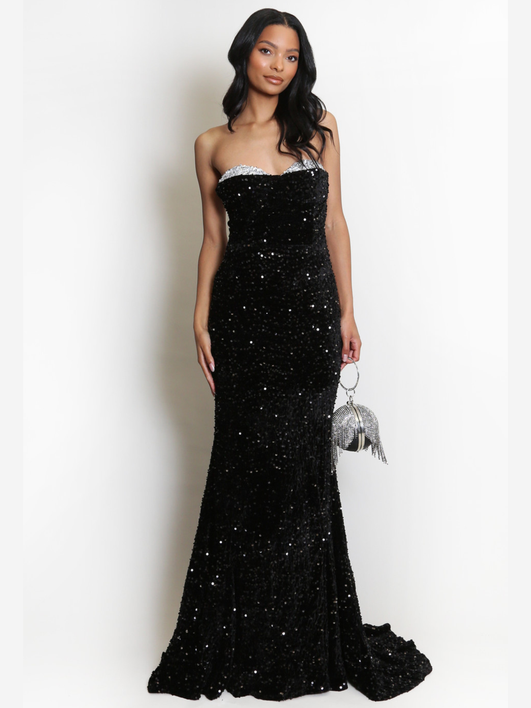 Full length shot. Model stands before a plain background. The model wears a black sequin maxi dress, with a fish tail hem and silver sequin sweetheart neckline. The model has light brown skin and long black hair. The model faces the camera, she holds a Diamante hand bag, the fish tail can be seen. 