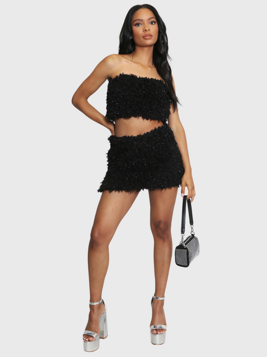 Full length shot. Woman wearing a black textured two-piece outfit on a white background. The co-ordinated set has a shaggy texture with silver flex detailing, the shaggy bandeau crop top has a matching mini skirt. The model has light brown skin and long black hair, she faces the camera. The model wears silver platform heels, placing one hand on her hip and the other by her side, holding a hand bag. 