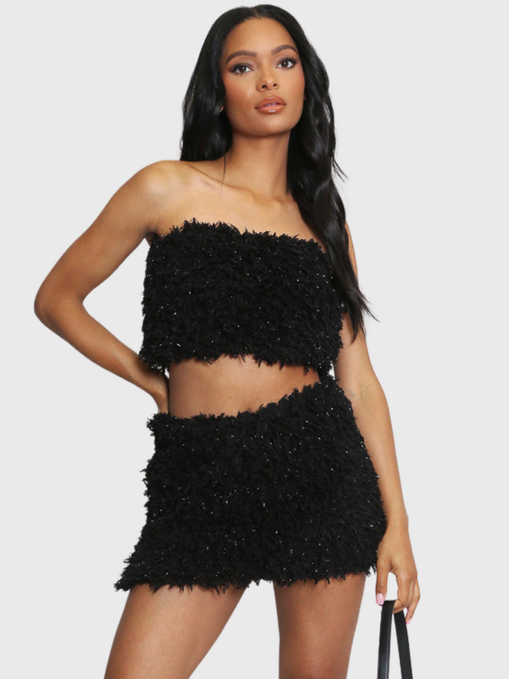 Woman wearing a black textured two-piece outfit on a white background. The co-ordinated set has a shaggy texture with silver flex detailing, the shaggy bandeau crop top has a matching mini skirt. The model has light brown skin and long black hair, she faces the camera and places  one hand on her hip and the other by her side. 