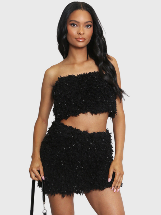 Woman wearing a black textured two-piece outfit on a white background. The co-ordinated set has a shaggy texture with silver flex detailing, the shaggy bandeau crop top has a matching mini skirt. The model has light brown skin and long black hair, she faces the camera and places both hands by her sides. 