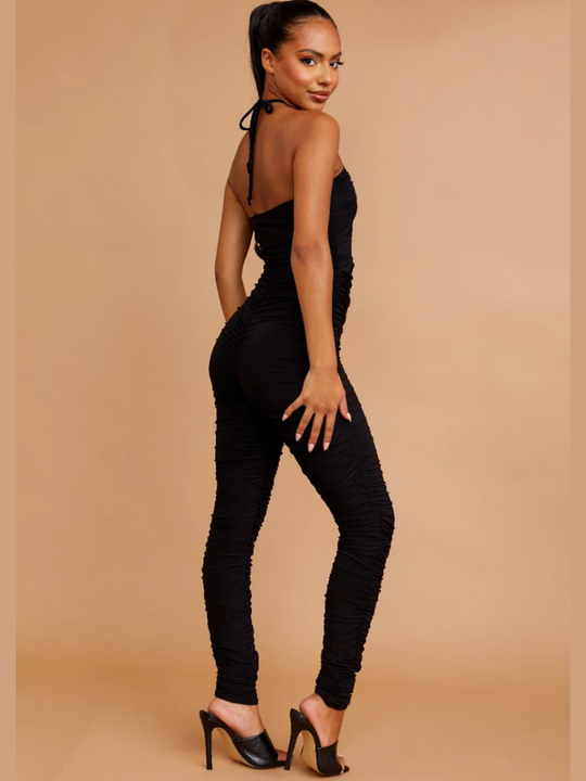 Model wears a black slinky ruched jumpuit with cut out front and adjustable criss cross halter neck fastening, which can be worn in a criss cross or traditional fastening. Model stands to the side and places her hands on her hips and turns her head to the camera. Model wars black muled heels.