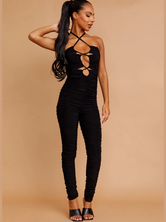 Full length shot. Model wears a black slinky ruched jumpuit with cut out front and adjustable criss cross halter neck fastening, which can be worn in a criss cross or traditional fastening. Model has light brown skin and wears her her hair slicked back with a long ponytail.  She places one hand on her head and the other by her thigh. The model wears black muled high heel shoes.  