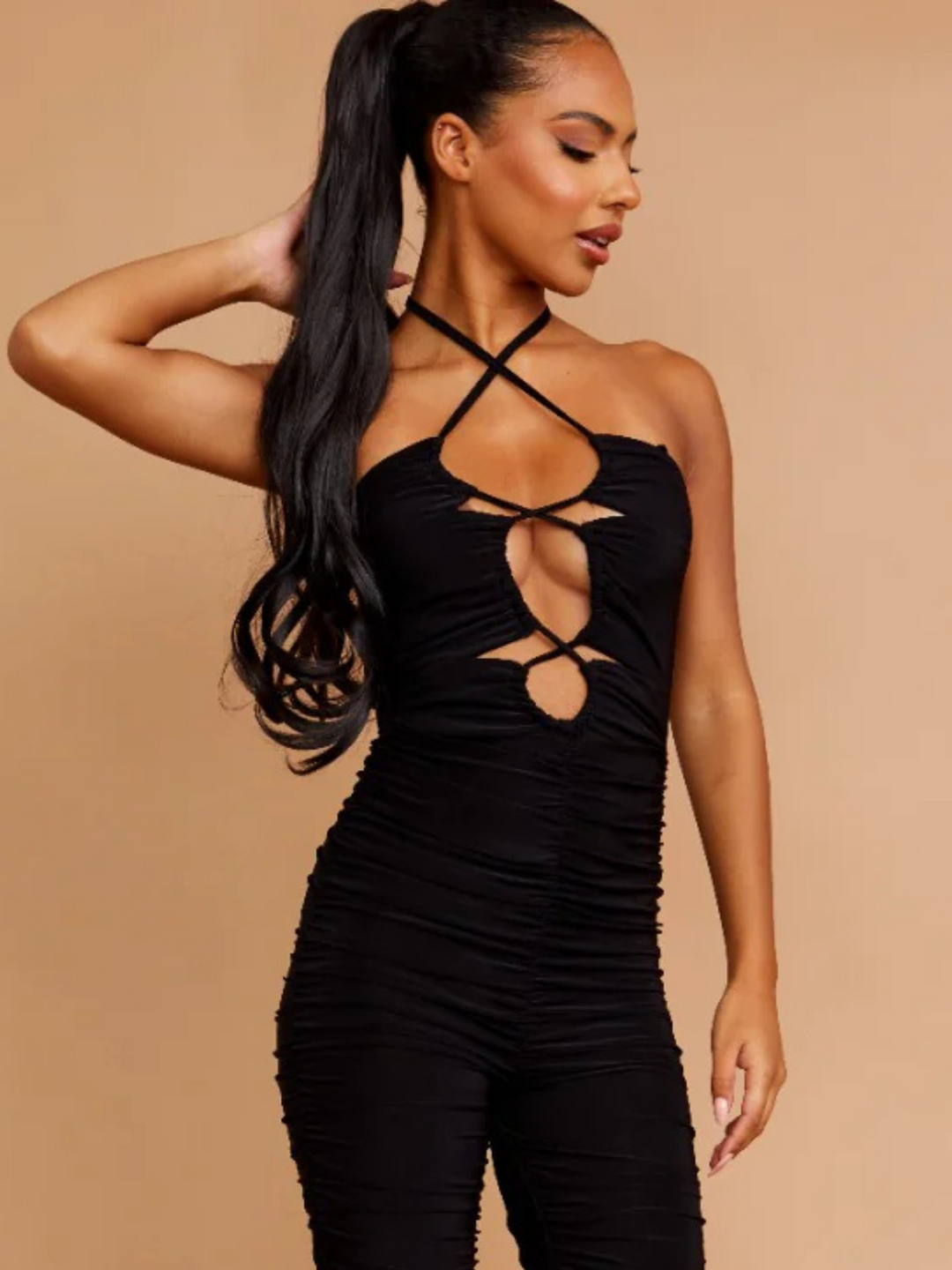 Model wears a black slinky ruched jumpuit with cut out front and adjustable criss cross halter neck fastening, which can be worn in a criss cross or traditional fastening. Model has light brown skin, she has her hair slicked back hair with a long wavy pony tail. Model stands with one hand behind her head and the other by her side.