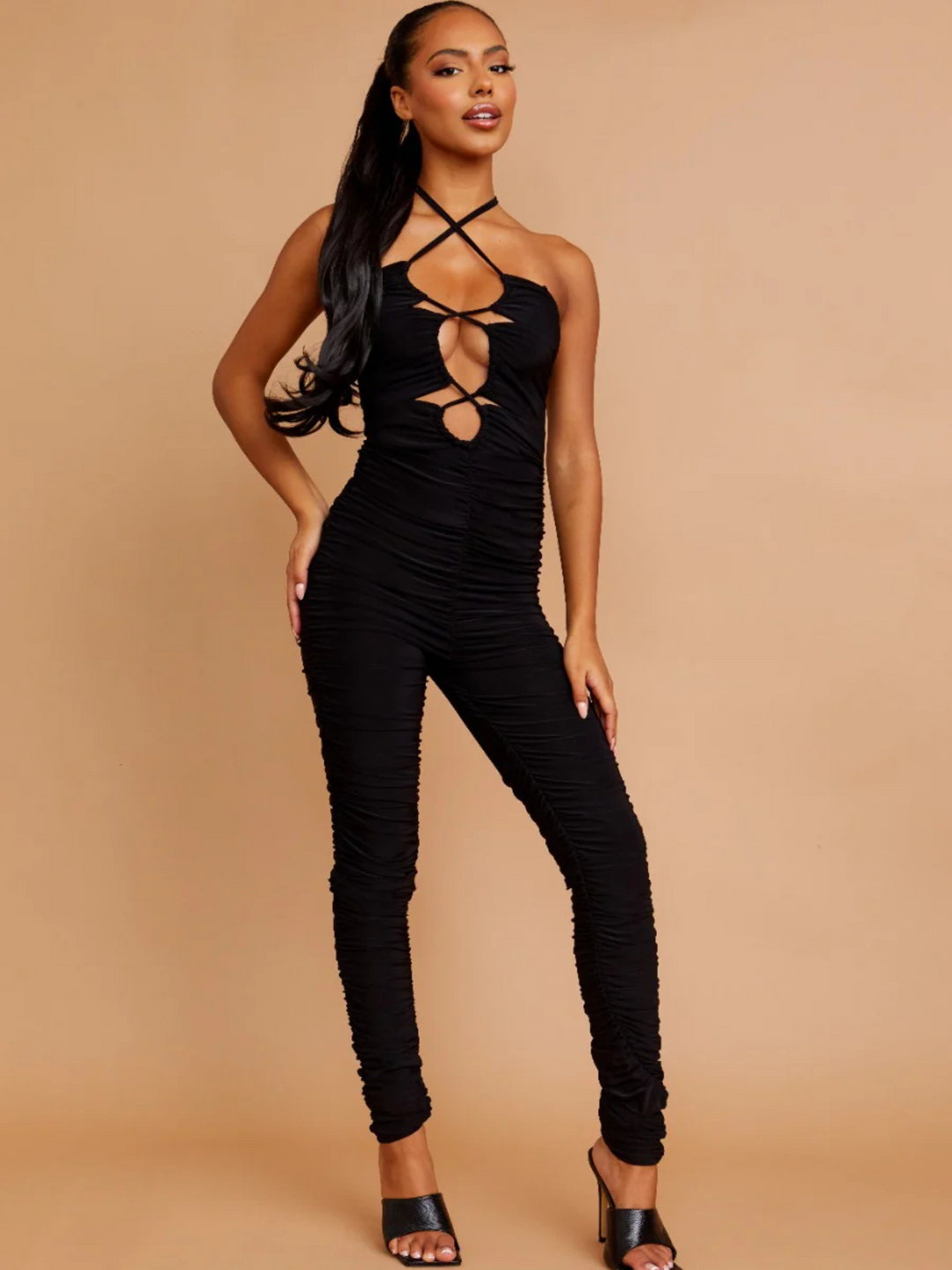 Full length shot. Model wears a black slinky ruched jumpuit with cut out front and adjustable criss cross halter neck fastening, which can be worn in a criss cross or traditional fastening. Model has light brown skin and wears her her hair slicked back with a long ponytail.  She places one hand on her thigh  and the other on her leg.