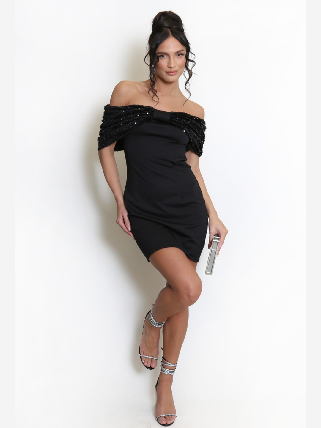 Full length shot. Model stands before a light grey background, she wears a black special occasion mini dress with a bardot off the shoulder sequin bow design. The model has tanned skin and black hair, she raises one leg above the ground and holds a silver clutch.