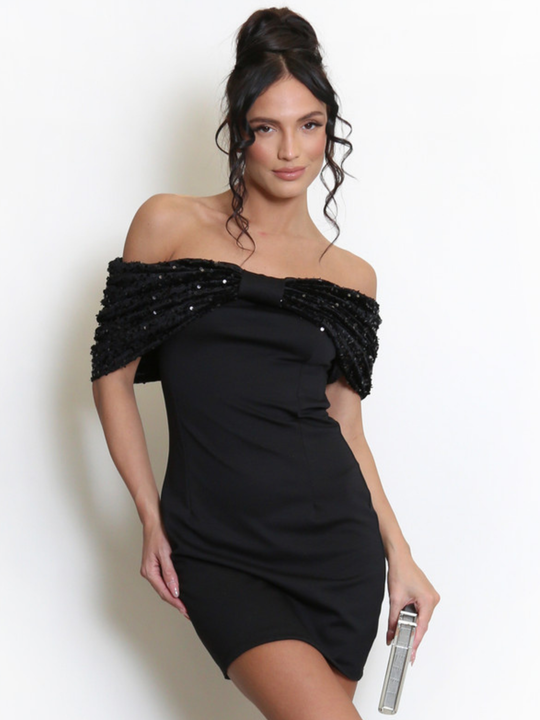 Model stands before a light grey background, she wears a black special occasion mini dress with a bardot off the shoulder sequin bow design. The model has tanned skin and black hair, she faces the camera and holds a clutch in one hand. 