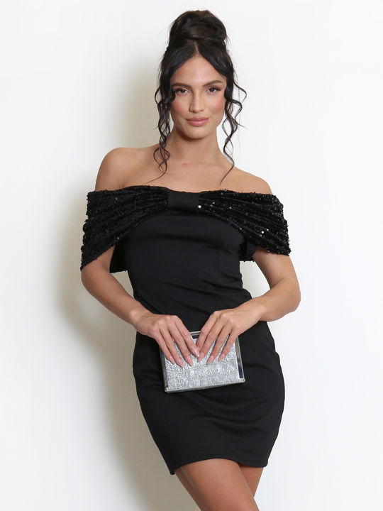 Model stands before a light grey background, she wears a black special occasion mini dress with a bardot off the shoulder sequin bow design. The model has tanned skin and black hair, she faces the camera and holds a silver clutch. The front of the dress can be seen, including the the bardot off the shoulder black sequin design.