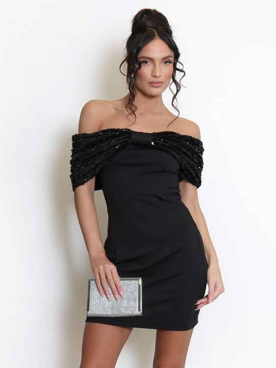 Model stands before a light grey background, she wears a black special occasion mini dress with a bardot off the shoulder sequin bow design. The model has tanned skin and black hair, she faces the camera and holds a clutch in one hand and the other by her side. The black front sequin bow can seen.