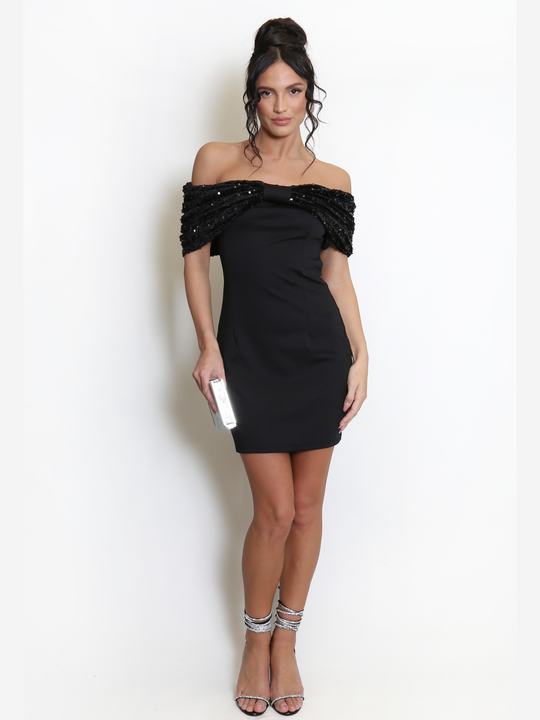 Full length shot. Model stands before a light grey background, she wears a black special occasion mini dress with a bardot off the shoulder sequin bow design. The model has tanned skin and black hair, she faces forward and has her hands by her sides. The front of the sequinned off the shoulder sleeves can be seen. The model wears lace up diamanté stiletto heels. 