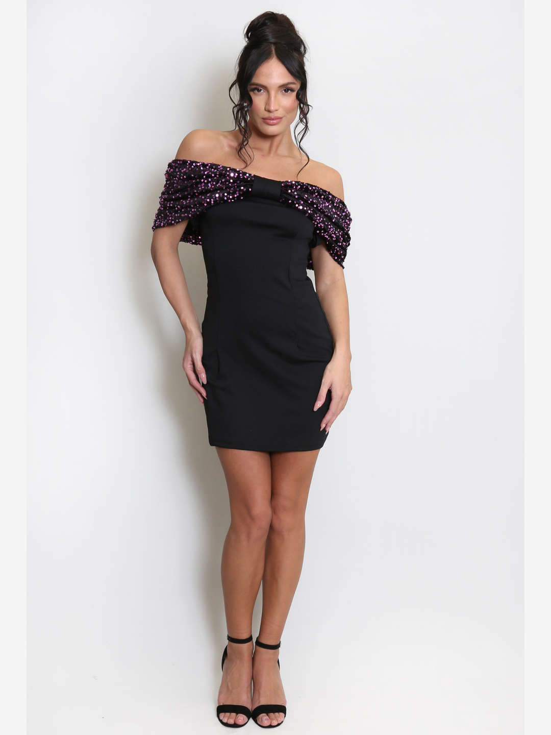 Full length shot. Model stands before a light grey background, she wears a black special occasion mini dress with a purple sequin bardot off the shoulder design. The model has tanned skin and black hair, she faces the camera and wears black heels.