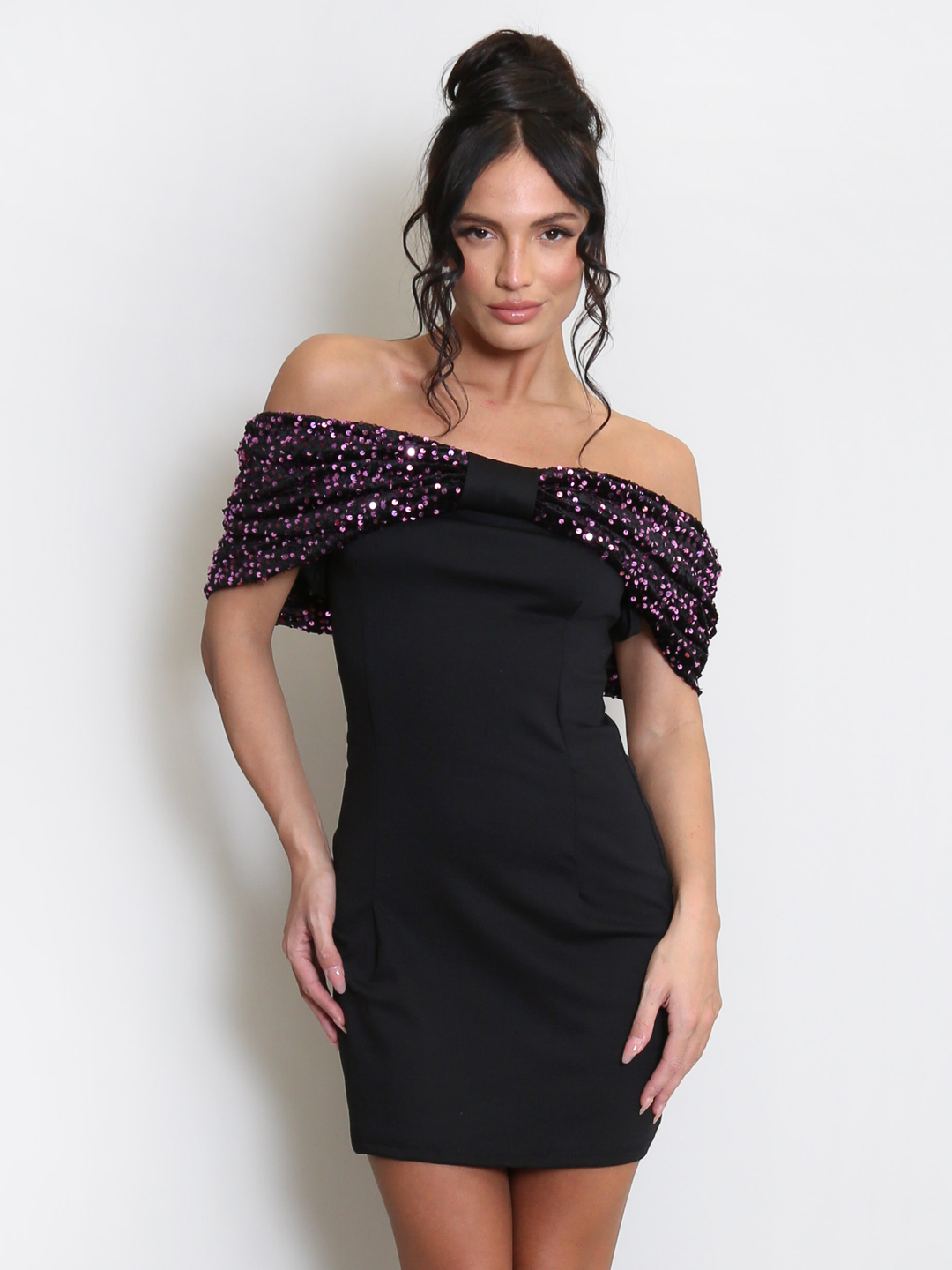 Model stands before a light grey background, she wears a black special occasion mini dress with a purple sequin bardot off the shoulder design. The model has tanned skin and black hair, she stands with both hands by her sides and looks directly into the camera. 