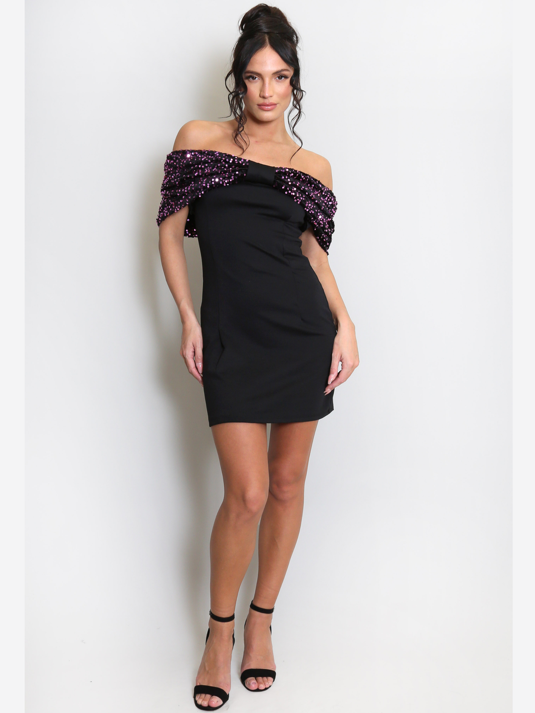 Full length shot. Model stands before a light grey background, she wears a black special occasion mini dress with a purple sequin bardot off the shoulder design. The model has tanned skin and black hair, she stands with one hand in front of the other and places both hands by her sides. 
