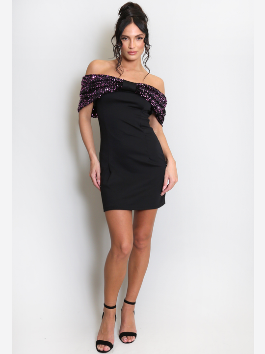Full length shot. Model stands before a light grey background, she wears a black special occasion mini dress with a purple sequin bardot off the shoulder design. The model has tanned skin and black hair, she stands with one hand in front of the other and places both hands by her sides. 
