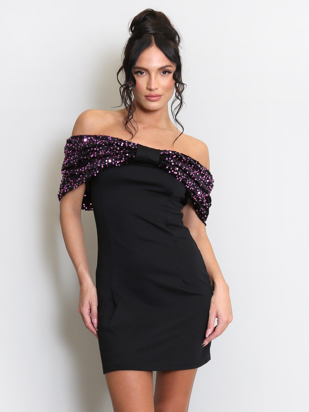 Model stands before a light grey background, she wears a black special occasion mini dress with a purple sequin bardot off the shoulder design. The model has tanned skin and black hair, she faces the camera and places her hands by her sides., the front of the dress can be seen including including the purple sequin sleeves.