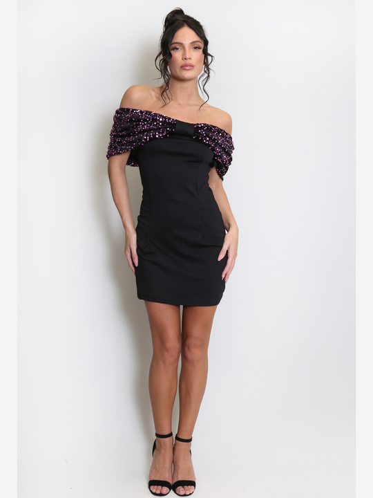 Full length shot. Model stands before a light grey background, she wears a black special occasion mini dress with a purple sequin bardot off the shoulder design. The model has tanned skin and black hair, she places both hands by her sides and wears a black heels. 