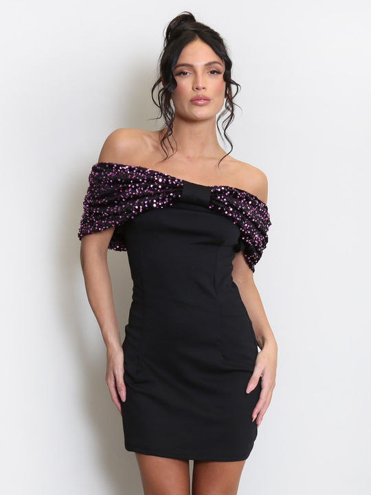 Model stands before a light grey background, she wears a black special occasion mini dress with a purple sequin bardot off the shoulder design. The model has tanned skin and black hair, she faces the camera and places both hands by her sides. 