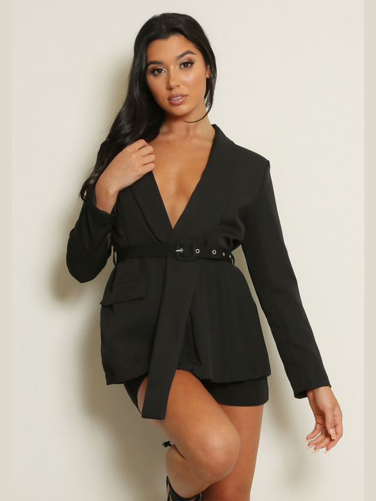 Model wears a black blazer with a v shaped cut. The black blazer has a pleated trim design and a belted adjustable waist. The model looks directly into the camera and  has tanned skin and black hair; she leans on the wall and bends one knee, has one hand by her side and the other touches the edge of the lapel. 