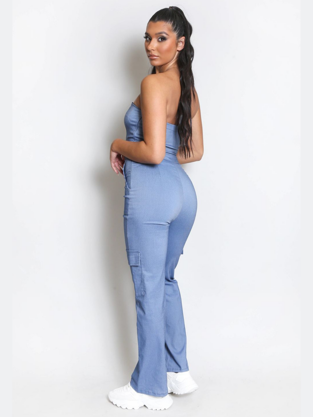 Full length shot. Female model wears a blue cargo jumpsuit that has cargo pockets, is strapless and has a bandeau neckline. The model has tanned skin and long black hair,  she stands to the side, the back and side of the jumpsuit can be seen, including the thigh cargo pockets. The model wears white chunky trainers.