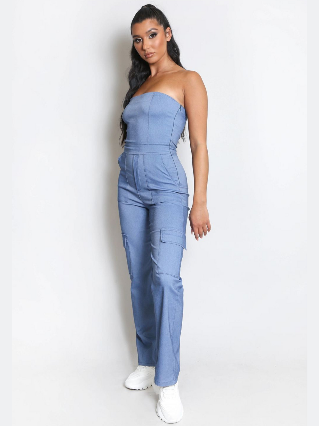 Full length shot. Female model wears a blue cargo jumpsuit that has cargo pockets, is strapless and has a bandeau neckline. The model has tanned skin and long black hair, she faces the camera placing one leg in the front of the other, the cargo pockets can be seen. The model wears white chubky trainers. 