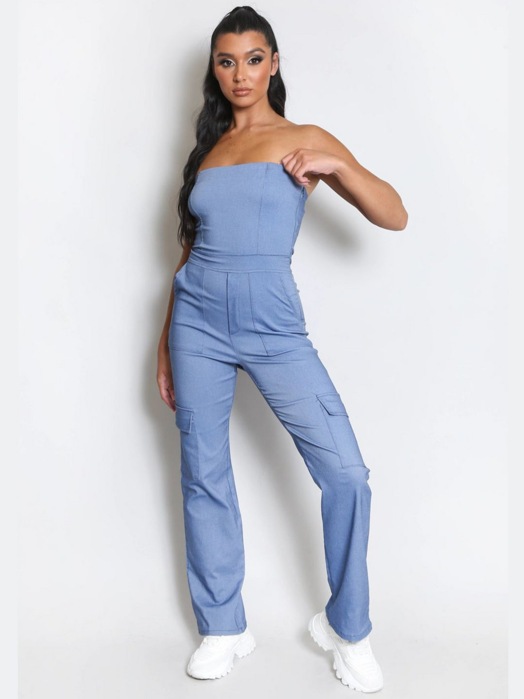 Full length shot. Female model wears a blue cargo jumpsuit that has cargo pockets, is strapless and has a bandeau neckline. The model has tanned skin and long black hair, she faces the camera and places one hand by her side and the other at her neckline. The Model wears chunky white trainers. 