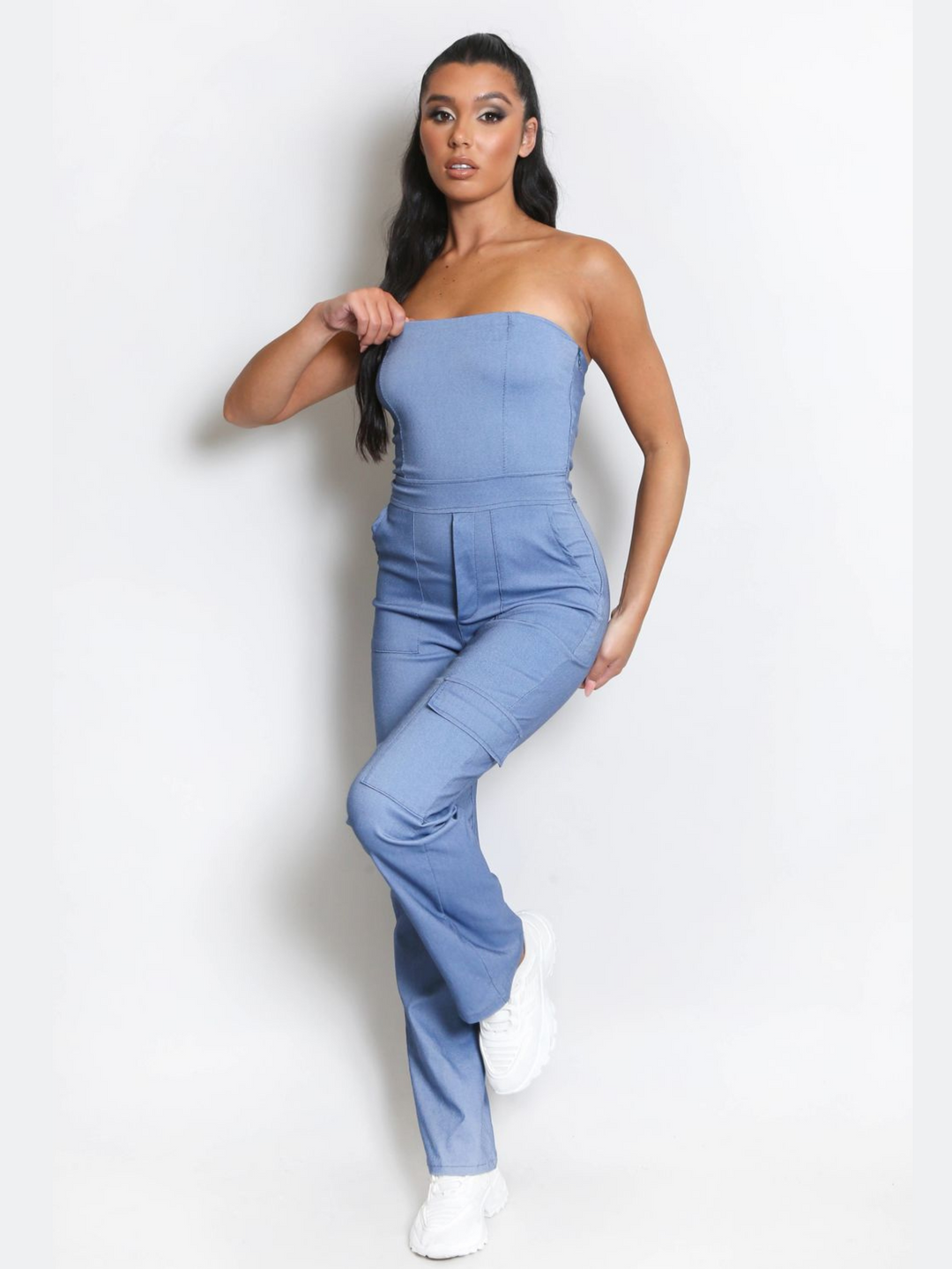 Full length shot. Female model wears a blue cargo jumpsuit that has cargo pockets, is strapless and has a bandeau neckline. The model has tanned skin and long black hair, she faces the camera,with one leg raised. The model places one hand by her side and the other on her neckline. The model wears white chunky trainers. 