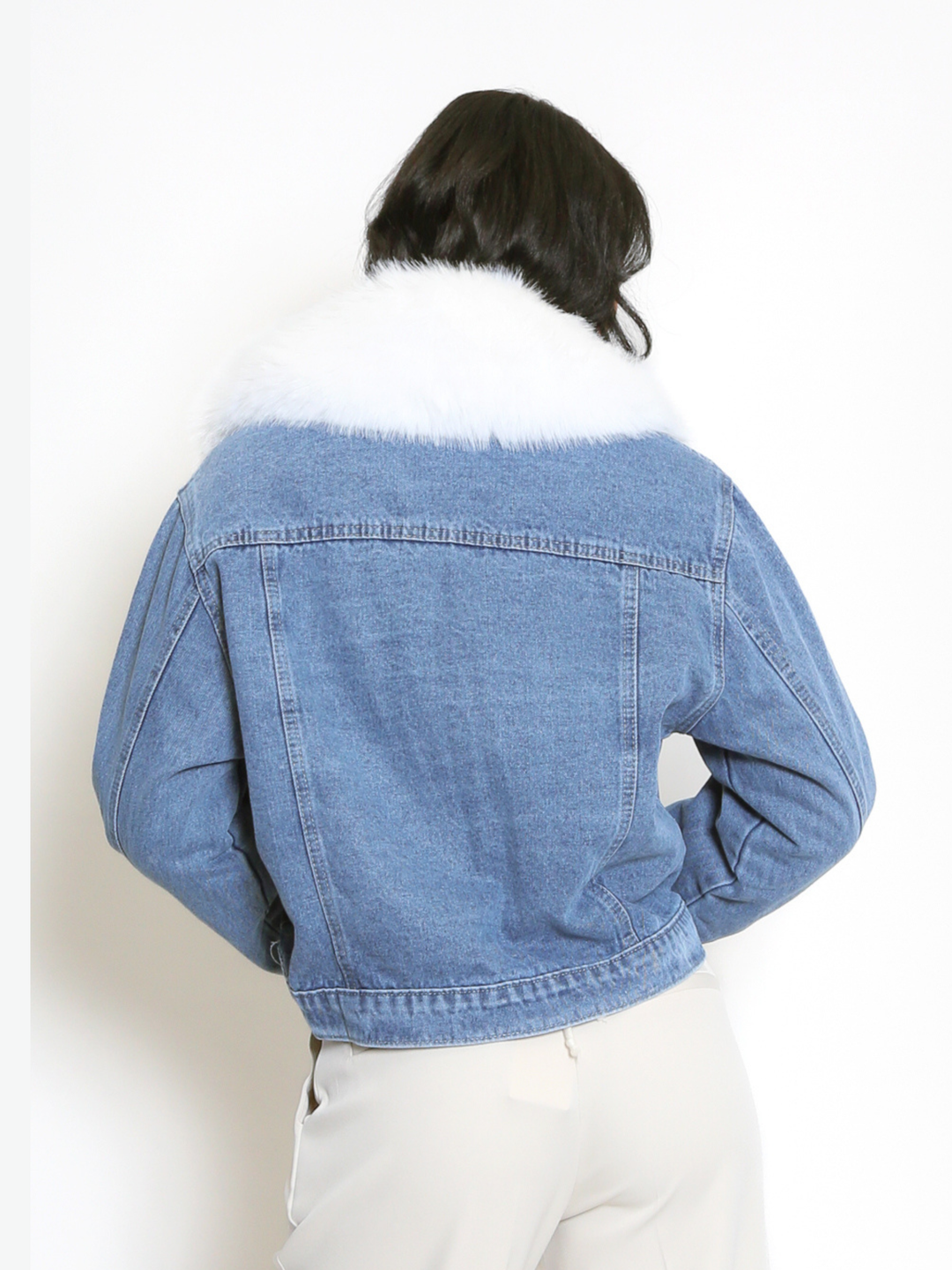 Female model stands in front of a plain background. The model wears a blue denim jacket with white faux fur collared trim, white front faux fur trimming and white faux fur cuffs on the long denim sleeves. The model has light brown skin and long black hair, she stands with her back to the camera, the back of the jacket can be seen, including the white faux fur collar. 