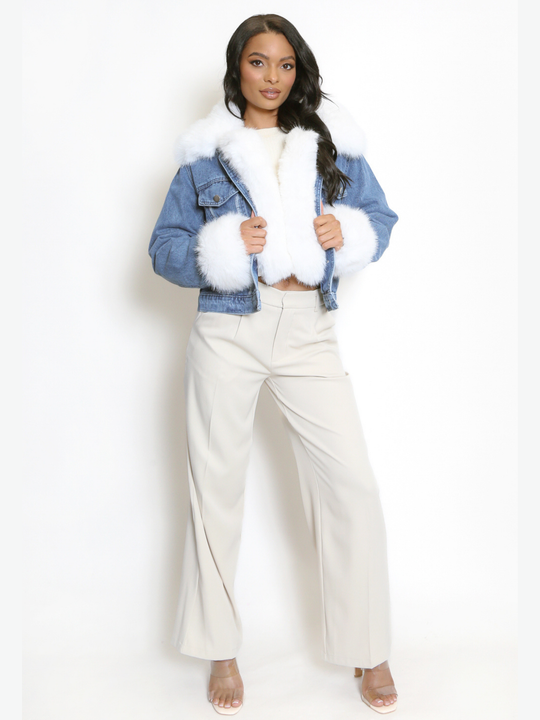 Full length shot. Female model stands in front of a plain background. The model wears a blue denim jacket with white faux fur collared trim, white front faux fur trimming and white faux fur cuffs on the long denim sleeves. The model has light brown skin and long black hair, she stands facing the camera with her hands pressed towards the centre of the  front jacket, she stands with one leg slightly in front of the other, she wears perspex heels and tanned trousers. 