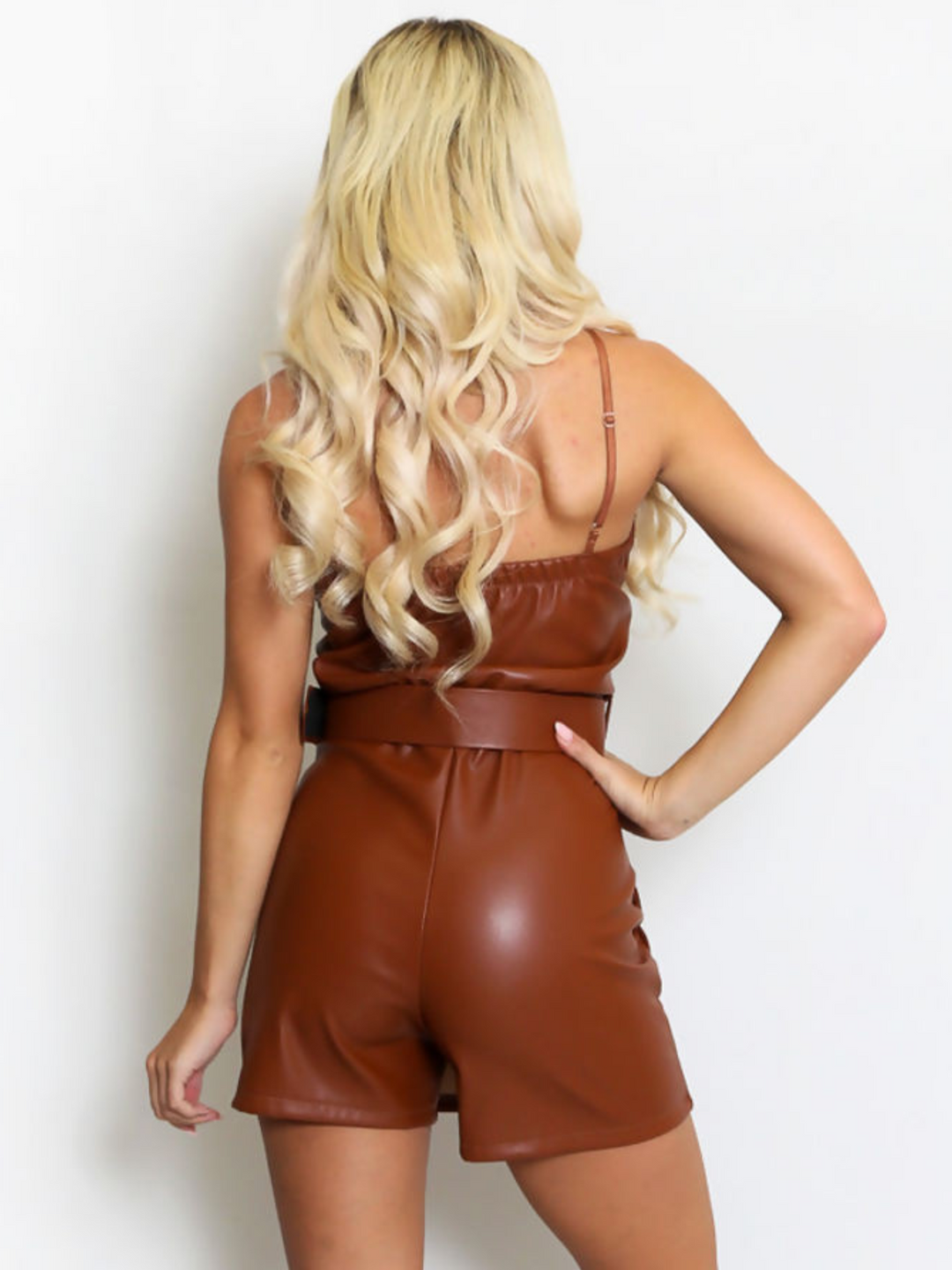 Model wears a PU leather playsuit that has a dress skort effect, the front of the dress appears to be a mini dress, but from behind it is clear that it is a playsuit, as the shorts is visible. The model has tanned skin and blonde hair, she stands with her back to the camera, the back of the playsuit can be seen, including the shorts and the back of the adjustable belt. She stands with one hand on her hip and the the other by her side. 