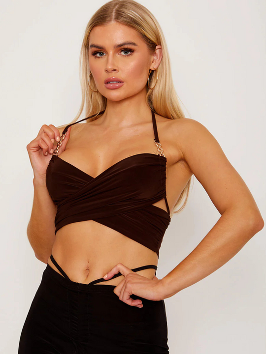 This image showcases a striking brown wrap-style crop top, featuring an alluring halter neck and sophisticated gold chain detailing on the straps. The crop top's design is both elegant and bold, with a snug fit that accentuates the silhouette, making it a standout piece for evening wear or special occasions. Model has tanned skin, she places one hand on the gold strap detailing and the other on her hip. 