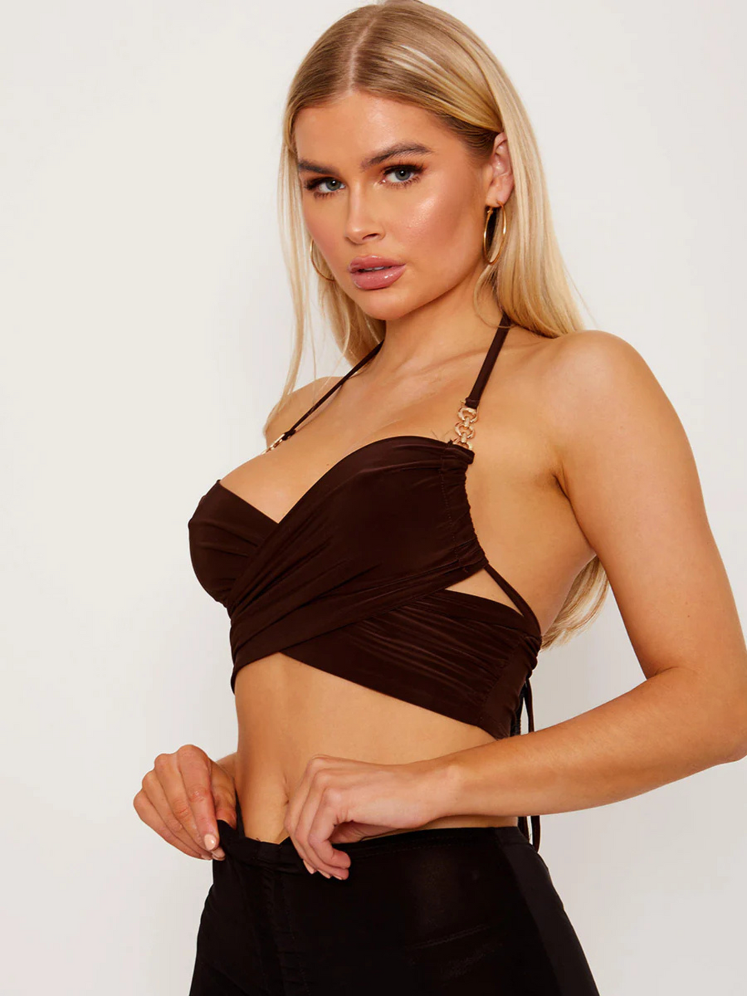 Model wears a brown wrap style crop with tie back fastening.The slinky strap has gold chain accents. Model has tanned skin and blonde hair,  she stands slightly to the side and places both hands on her trousers, she turns her head slightly to look directly into the camera.