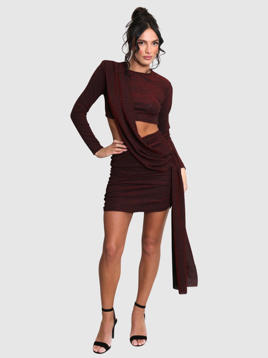 Full length shot. Model stands in front of a plain background. The model wears a cut out lurex burgandy mini dress, with long sleeves and diagnal draped front and rounded neck. The model has tanned skin and black hair that is in a high bun, she faces the camera with one hand on his hip and the other by her side, she wears a pair of strappy balck heels. 