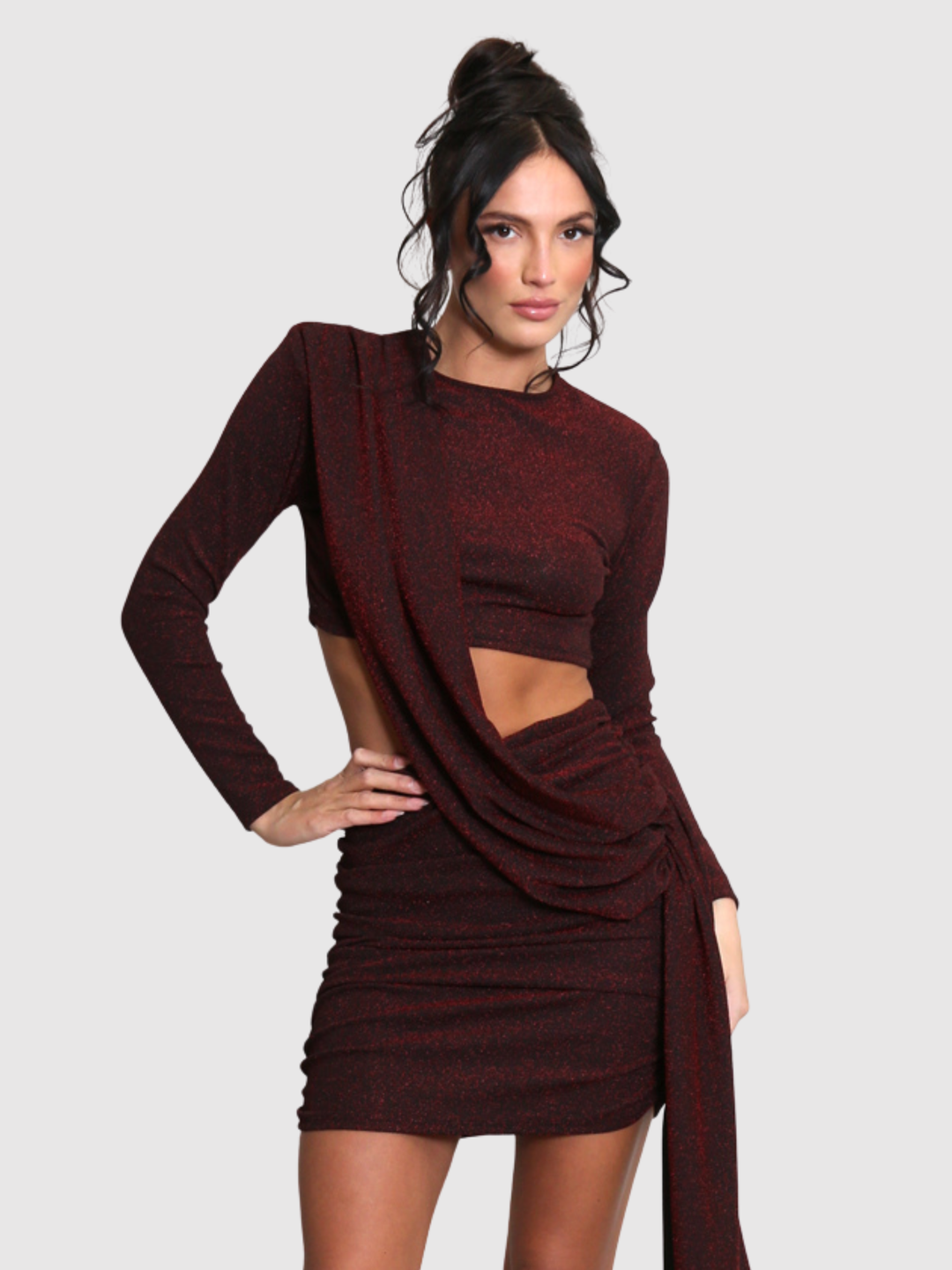 Model stands in front of a plain background. The model wears a cut out lurex burgandy mini dress, with long sleeves and diagnal draped front and rounded neck. The model has tanned skin and black hair that is in a high bun. The model places one hand on her hip and the other by her side.