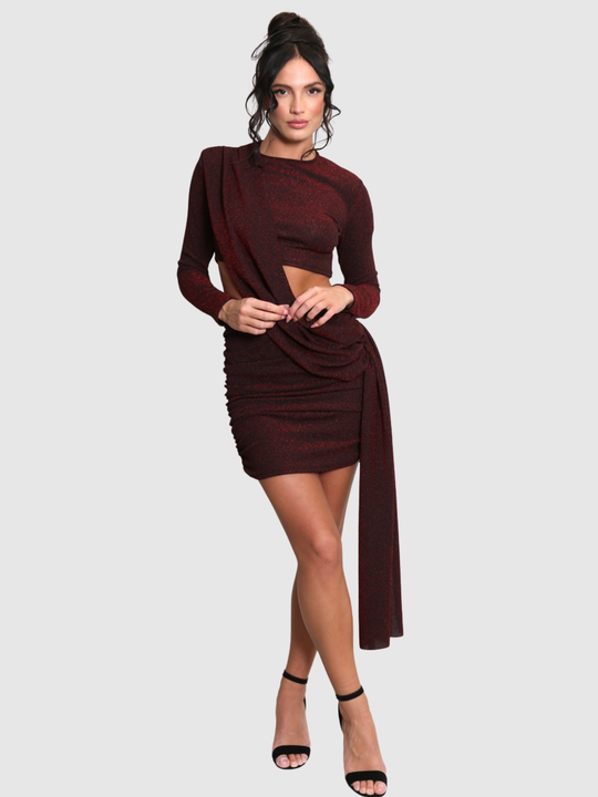 Full length shot. Model stands in front of a plain background. The model wears a cut out lurex burgandy mini dress, with long sleeves and diagnal draped front and rounded neck. The model has tanned skin and black hair that is in a high bun, she crosses her legs placing both hands in front of her stomach and looks directly into the camera. 
